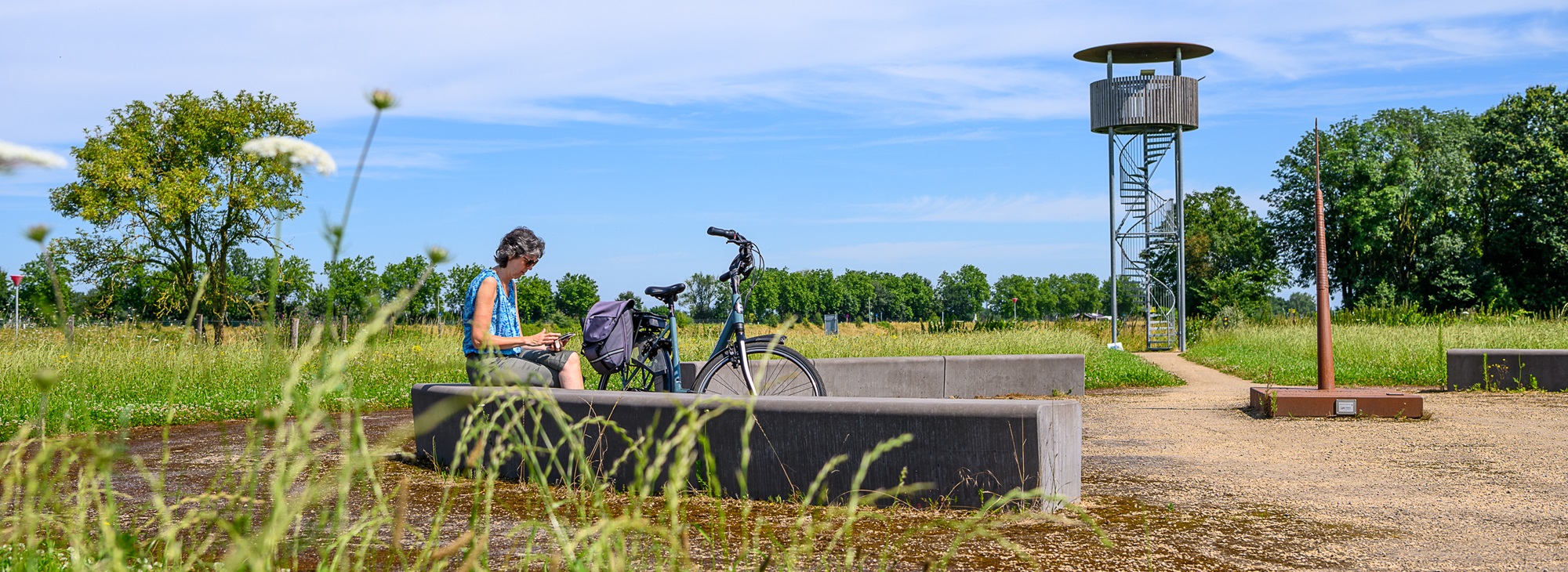 Vrouw met fiets rust uit bij een zitplek in een groen landschap met uitkijktoren op de achtergrond, onder een blauwe lucht in Stevensweert