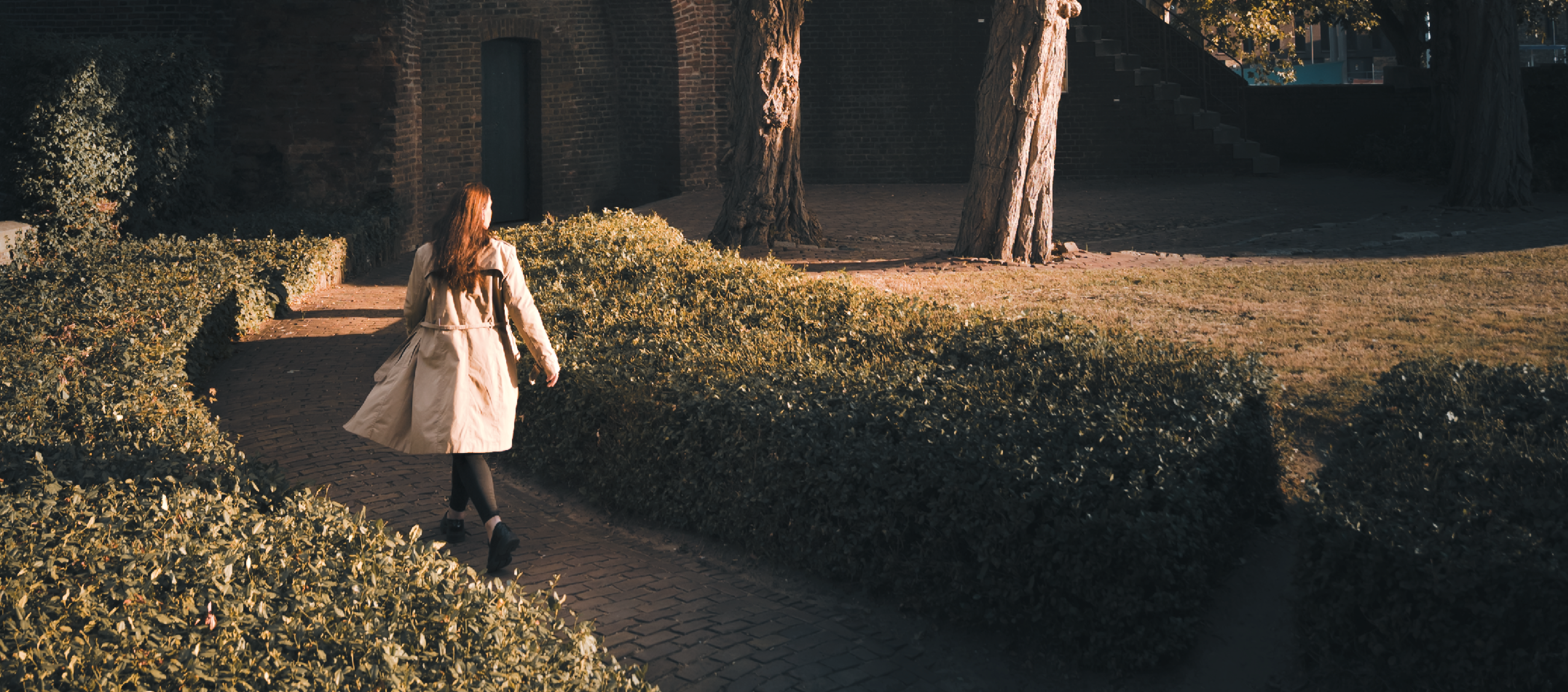 Vrouw in een lichte jas loopt door een tuinpad langs een oude stadsmuur met bomen in het zonlicht.