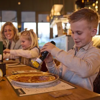 Jongen schenkt stroop over zijn pannenkoek bij restaurant La Selva op Recreatiepark de Leistert, met zijn gezin op de achtergrond.